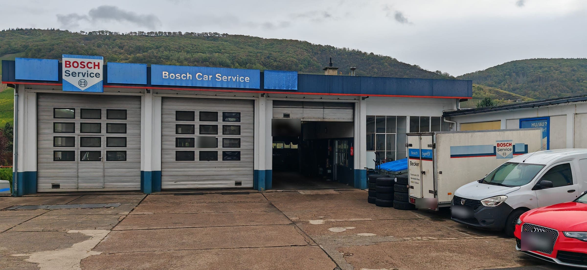 Die Werkstatthalle von vorn: drei Tore unter dem Bosch-Car-Service-Schild, eines offen, rechts der Anhänger des Betriebs, dahinter bewaldete Hänge