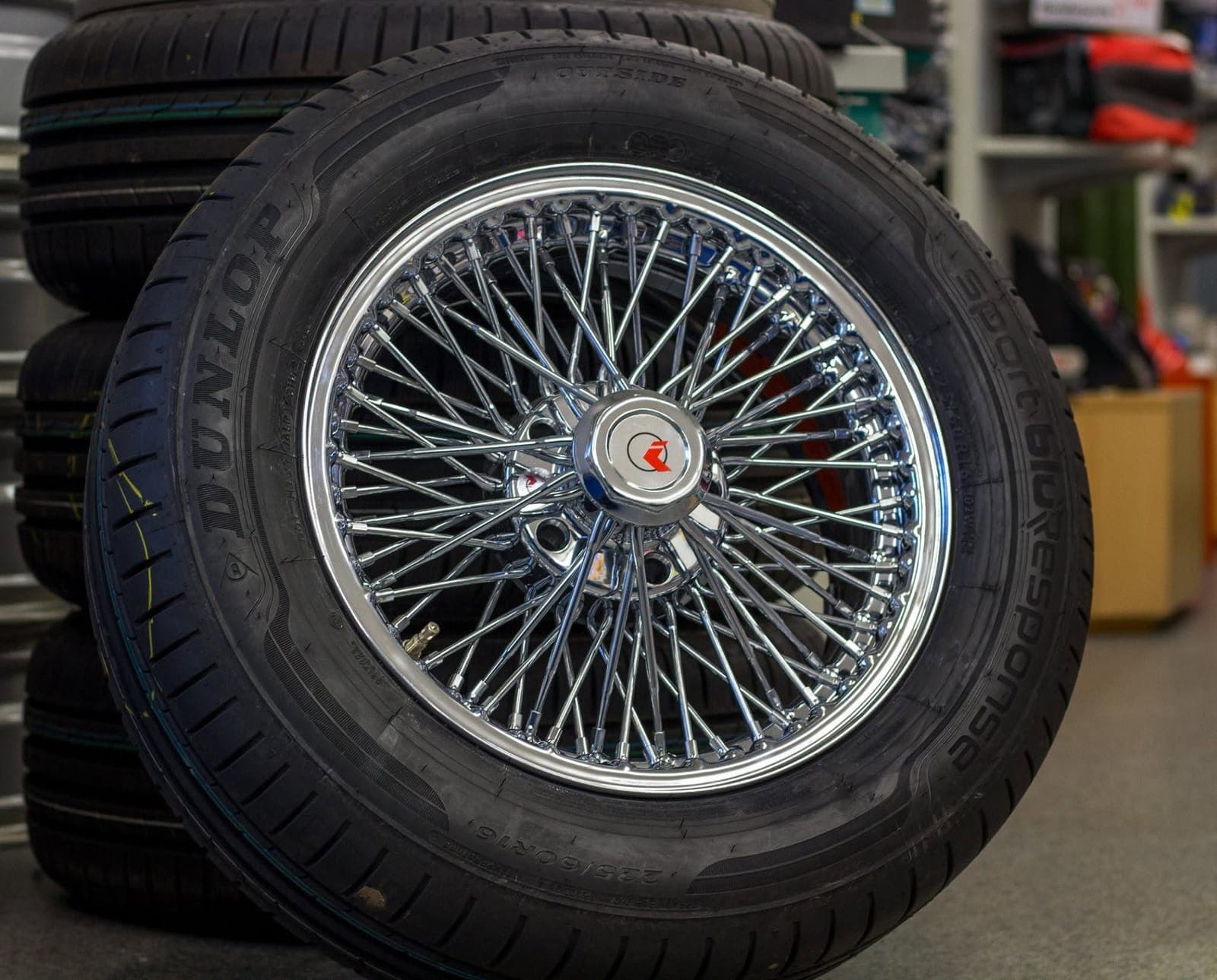 Verchromtes Speichenrad mit Zentralverschluss für einen Jaguar, darauf ein neuer Dunlop-Reifen, im Hintergrund weitere Reifen