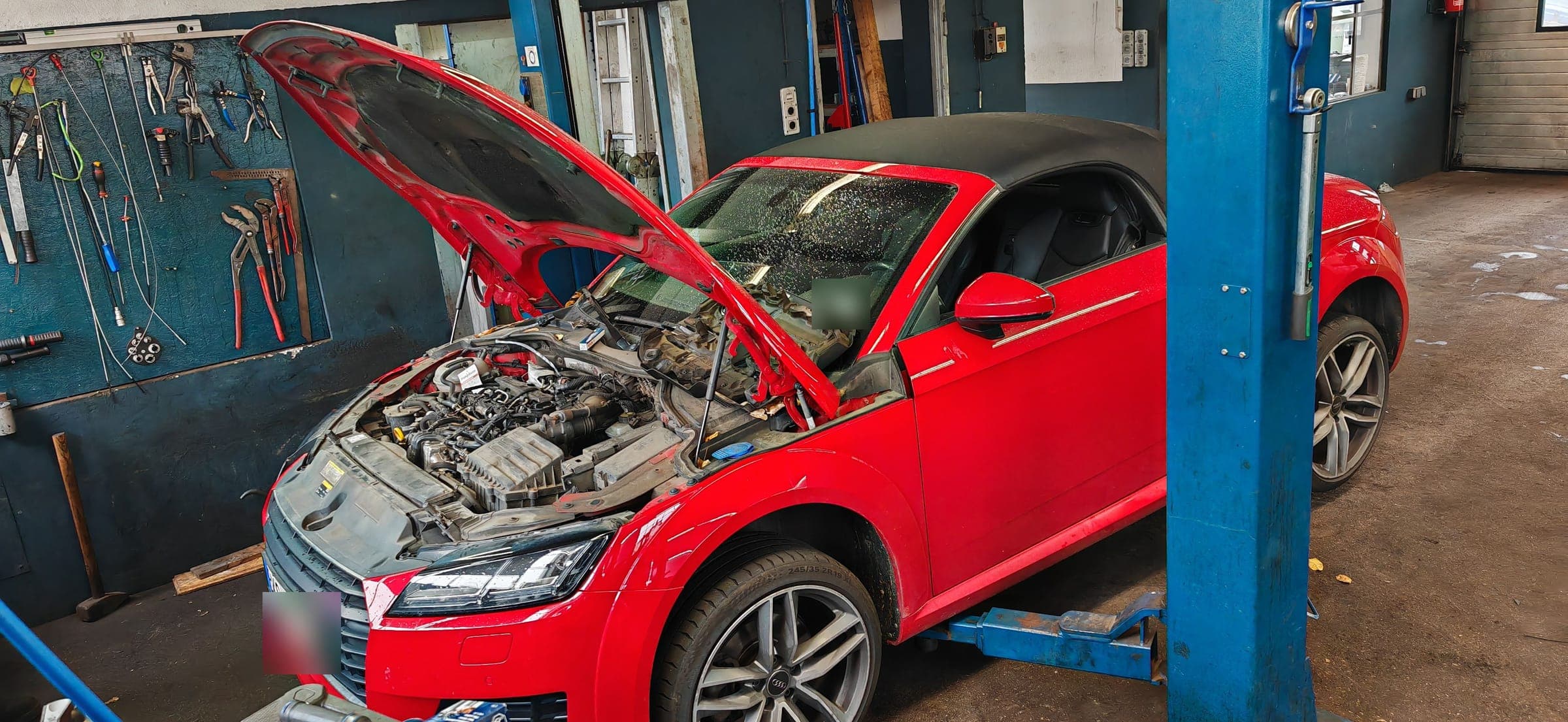 Ein roter Audi TT mit geöffneter Motorhaube neben der Hebebühne, links die Werkzeugwand