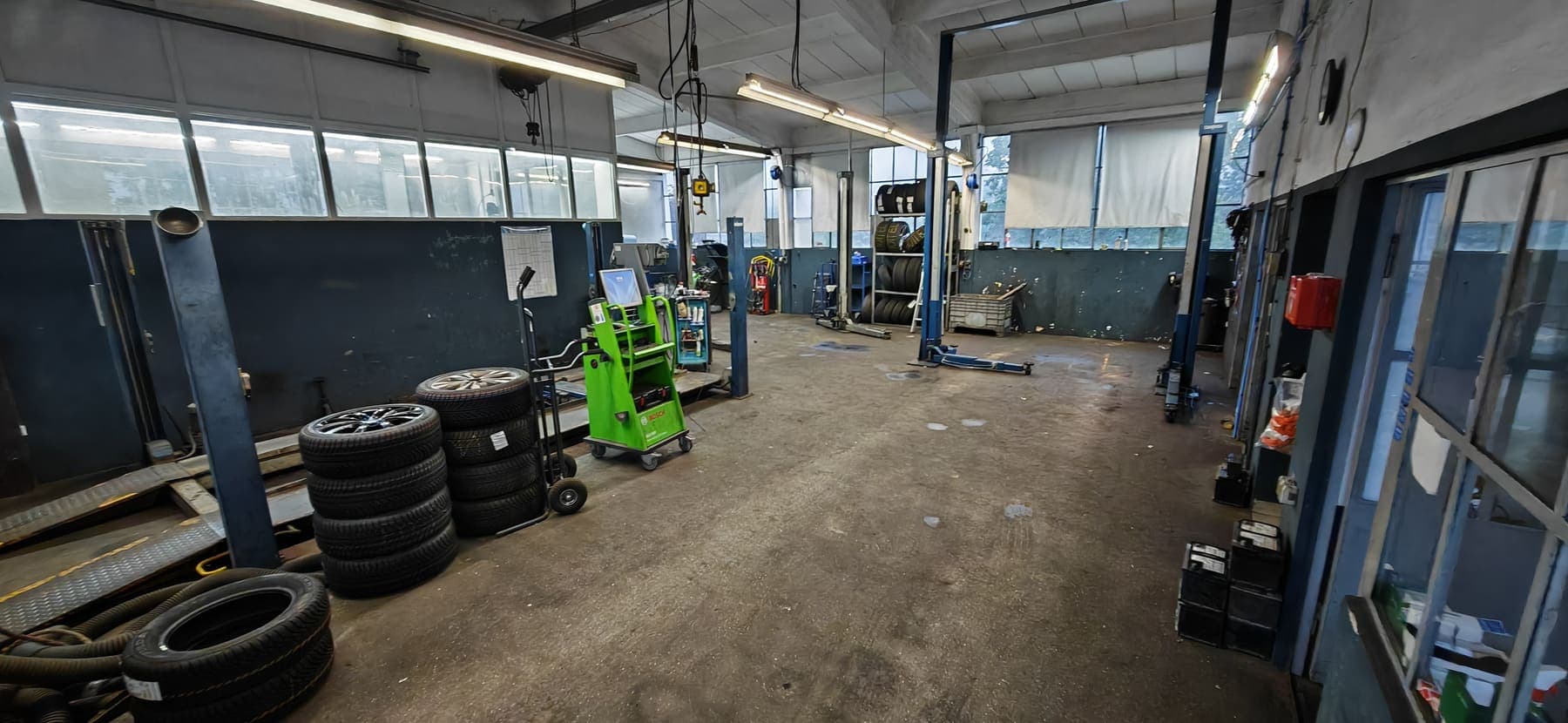 Blick in die Werkstatthalle: zwei Zwei-Säulen-Hebebühnen, links die abgesenkte Auffahrrampe einer Vier-Säulen-Bühne, davor ein Stapel Kompletträder, ein Bosch-Diagnosewagen und ein Reifenregal an der Rückwand. Über der Trennwand Lichtschächte, dahinter eine weitere Box.